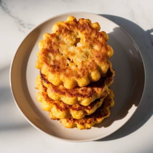 A stack of round, Southern-style corn fritters with a crispy crust sits on a beige plate, placed on a white marble surface under strong natural lighting.