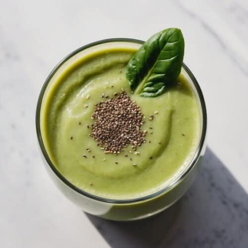 A breakfast smoothie featuring spinach in a glass, topped with chia seeds and a single basil leaf, sits elegantly on a marble surface.