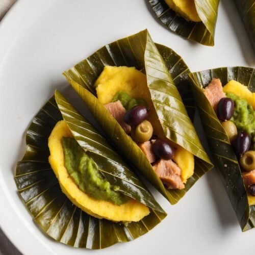 Three cornmeal dough parcels, reminiscent of Puerto Rican pasteles, are wrapped in leaves and filled with green sauce, olives, and a protein. These masa delights are elegantly arranged on a white plate with a cloth beside it.